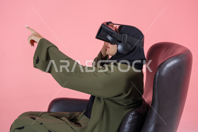 Technical and technological development, fun and entertainment watching through virtual reality glasses, a side portrait of a veiled Emirati Arab Gulf woman wearing a green abaya, wearing VR glasses, sitting on a comfortable chair and raising her hand in integration, enjoyment and spending recreational time playing video games, pink background