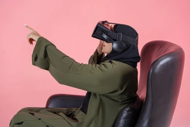 التطور التقني والتكنولوجي, المتعة و التسلية بالمشاهدة عبر نظارة الواقع الافتراضي، بورتريه من الجانب لامرأة اماراتية عربية خليجية محجبة ترتدي عباءة خضراء تضع نظارة الفي ار و تجلس على كرسي مريح و ترفع يدها باندماج, الاستمتاع و قضاء اوقات ترفيهية في اللعب بالعاب الفيديو، خلفية وردية