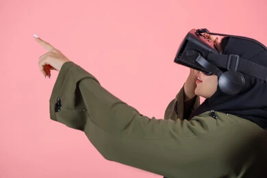 Watching 3D VR movies, interacting and simulating the imaginary virtual world, enjoying and spending leisure time playing video games, modern technologies and technological devices, a close-up portrait of a veiled Emirati Arab Gulf woman wearing a green abaya and pointing her hand upwards in a blended manner, pink background