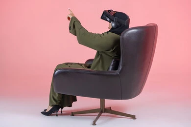 Fun and entertainment watching through virtual reality glasses, a side portrait of a veiled Emirati Arab Gulf woman wearing a green abaya, wearing VR glasses, sitting on a comfortable chair and raising her hand in immersion, enjoyment and spending recreational time playing video games, pink background
