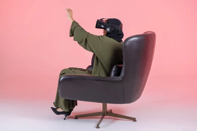 محاولة لمس شيء ما، تقنيات حديثة وأجهزة تكنولوجية, التفاعل و محاكاة العالم الافتراضي الخيالي, بورتريه من الجانب لامرأة اماراتية عربية خليجية محجبة ترتدي عباءة خضراء تضع نظارة الفي ار و تجلس على كرسي مريح و ترفع يدها باندماج, خلفية وردية