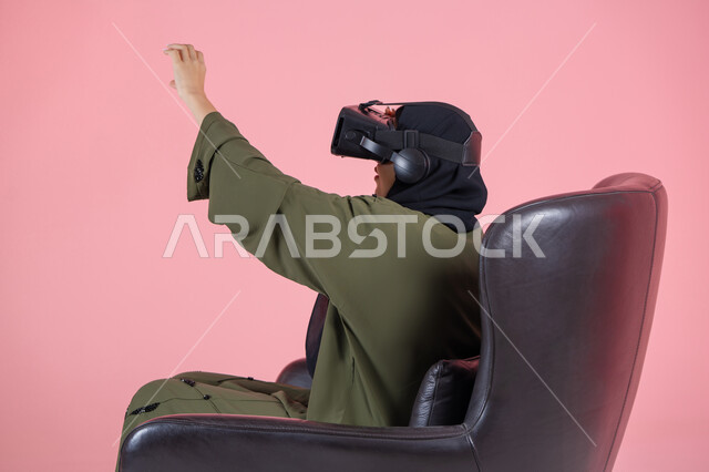 Enjoy and spend leisure time playing video games, modern technologies and technological devices, interaction and simulation of the imaginary virtual world, portrait from the back of a veiled Emirati Arab Gulf woman wearing a green abaya, wearing VR glasses and raising her hand in integration, pink background