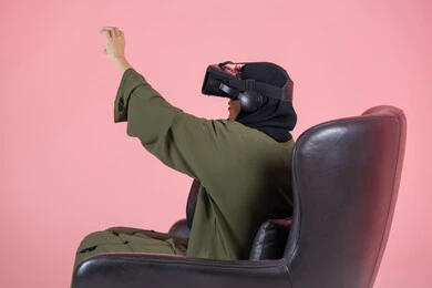 الاستمتاع و قضاء اوقات ترفيهية في اللعب بالعاب الفيديو، تقنيات حديثة وأجهزة تكنولوجية, التفاعل و محاكاة العالم الافتراضي الخيالي, بورتريه من الخلف لامرأة اماراتية عربية خليجية محجبة ترتدي عباءة خضراء و تضع نظارة الفي ار و ترفع يدها باندماج, خلفية وردية