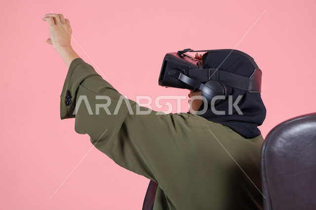 Interaction and simulation of the imaginary virtual world, enjoyment and spending leisure time playing video games, modern technologies and technological devices, close-up portrait of a veiled Emirati Arab Gulf woman wearing a green abaya, wearing VR glasses and raising her hand in integration, pink background