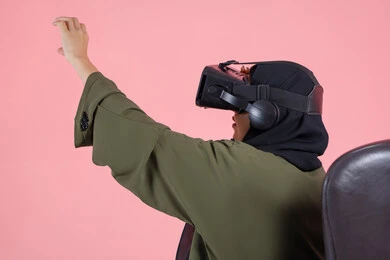 Interaction and simulation of the imaginary virtual world, enjoyment and spending leisure time playing video games, modern technologies and technological devices, close-up portrait of a veiled Emirati Arab Gulf woman wearing a green abaya, wearing VR glasses and raising her hand in integration, pink background