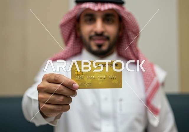 The possibility of paying bills remotely, a Gulf Arab Saudi man wearing traditional attire and a bisht uses a bank credit card for online shopping, purchasing, and payment through internet programs and applications.