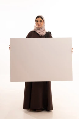 العروض والإعلانات التسويقية, لوحات توضيحية ووسائل تعليمية، بورتريه لطالبة عربية خليجية سعودية محجبة ترتدي العباءة تمسك بيديها لوحة بيضاء مستطيلة فارغة و تشير الية، النظر إلى الكاميرا بتعابير السعادة والسرور، موك اب مستطيل أبيض, خلفية بيضاء