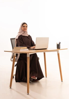 Looking at the camera with gestures of joy, modern technological devices in learning, following lectures through learning platforms, a portrait of a Saudi Gulf Arab student wearing a hijab and abaya sitting at a desk, doing homework and attending school lectures remotely via a tablet, white background.