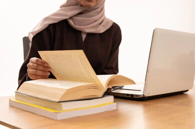 المحاضرات الجامعية عن بعد عبر الحاسب، كتابة  وتدوين الملاحظات خلال المحاضرة، الأجهزة التقنية الحديثة بالتعلم,، متابعة المحاضرات عن طريق منصات التعلم، بورتريه مقرب لطالبه عربيه خليجيه سعودية ترتدي الحجاب والعباءة تجلس على الطاولة المكتبيه ، خلفية بيضاء