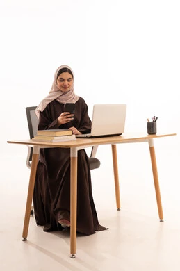 العمل على الهاتف المحمول خلال المحاضرة، المحاضرات الجامعية عن بعد عبر الحاسب، الأجهزة التقنية الحديثة بالتعلم,، متابعة المحاضرات عن طريق منصات التعلم، بورتريه مقرب لطالبه عربيه خليجيه سعودية ترتدي الحجاب والعباءة تجلس على الطاولة المكتبيه ، خلفية بيضاء