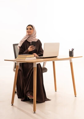 الهاتف المحمول خلال المحاضرة، المحاضرات الجامعية عن بعد عبر الحاسب، الأجهزة التقنية الحديثة بالتعلم,، متابعة المحاضرات عن طريق منصات التعلم، بورتريه مقرب لطالبه عربيه خليجيه سعودية ترتدي الحجاب والعباءة تجلس على الطاولة المكتبيه ، خلفية بيضاء