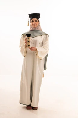 النجاح والتفوق والتخرج من الجامعة، إيماءات السعادة بالوصول الى الحلم، مناسبة واحتفال جامعي، الاحتفال بإنهاء الدراسة الجامعية، النظر الى الكاميرا بايماءات الفخر، بورتريه لخريجة عربية خليجية سعودية محجبة ترتدي زي التخرج والقبعة تحمل بيدها الشهادة، خلفية بيضاء