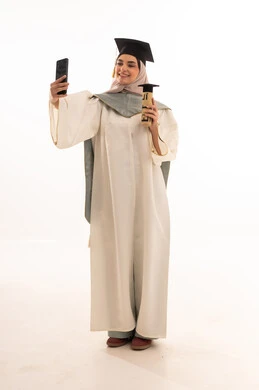 Taking a selfie on graduation day, gestures of happiness at reaching the dream, success and excellence and graduating from university, celebrating the completion of university studies, looking at the camera with gestures of pride, a portrait of a Saudi Arabian Gulf female graduate wearing graduation attire and cap holding her certificate in her hand, white background.
