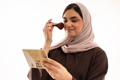 شابة سعودية تضع المكياج,  التزين والتجمل,  التسويق لمستحضرات ومنتجات التجميل, الاهتمام والعناية بالمظهر الخارجي, استخدام المساحيق التجميلية, بورتريه مقرب لامرأة عربية خليجية سعودية محجبة ترتدي العباءة السوداء تستعمل فرشاة أحمر الخدود, خلفية بيضاء