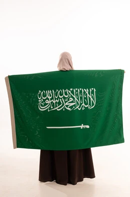 الاحتفال بالمناسبات الوطنية، الفخر و الاعتزاز بالانتماء للوطن، استذكار اليوم الوطني السعودي 23 سبتمبر، يوم العلم 11 مارس، رفع راية التوحيد والإسلام، بورتريه من الخلف  لامرأة عربية خليجية سعودية محجبة تحمل بيدها علم السعودية ، الجسم بالطول الكامل، خلفية بيضاء