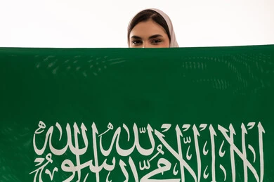 استذكار اليوم الوطني السعودي 23 سبتمبر، يوم العلم 11 مارس، الاحتفال بالأعياد والمناسبات الوطنية، رفع راية التوحيد والإسلام، بورتريه مقرب لامرأة عربية خليجية سعودية محجبة تحمل بيدها علم السعودية الاخضر، الفخر و الاعتزاز بالانتماء للوطن، خلفية بيضاء