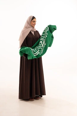 تقبيل العلم الوطني، الاحتفال بالمناسبات الوطنية، الفخر و الاعتزاز بالانتماء للوطن، استذكار اليوم الوطني السعودي 23 سبتمبر، يوم العلم 11 مارس، رفع راية التوحيد والإسلام، بورتريه لامرأة عربية خليجية سعودية محجبة تحمل بيدها علم السعودية ، الطول الكامل للجسم، خلفية بيضاء