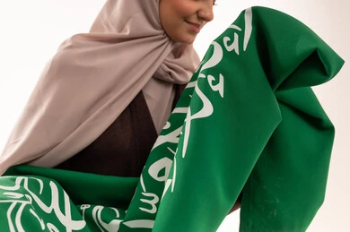 Commemoration of the Saudi National Day on September 23, Flag Day on March 11, celebration of national holidays and occasions, raising the banner of monotheism and Islam, a close-up portrait of a Saudi Gulf Arab woman in a hijab holding the green Saudi flag in her hand, pride and honor in belonging to the homeland, white background.