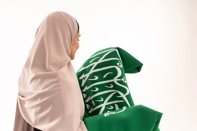 استذكار اليوم الوطني السعودي 23 سبتمبر، يوم العلم 11 مارس، الاحتفال بالأعياد والمناسبات الوطنية، رفع راية التوحيد والإسلام، بورتريه مقرب لامرأة عربية خليجية سعودية محجبة تحمل بيدها علم السعودية الاخضر، الفخر و الاعتزاز بالانتماء للوطن، خلفية بيضاء