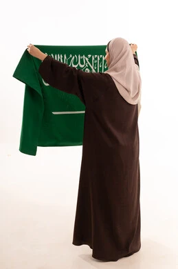 Celebrating national occasions, pride and honor in belonging to the homeland, commemorating Saudi National Day on September 23, Flag Day on March 11, raising the banner of monotheism and Islam, a portrait from behind of a Saudi Arabian Gulf woman wearing a hijab holding the Saudi flag in her hand, full-body, white background.