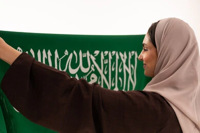 الاحتفال بالمناسبات الوطنية، الفخر و الاعتزاز بالانتماء للوطن، استذكار اليوم الوطني السعودي 23 سبتمبر، يوم العلم 11 مارس، رفع راية التوحيد والإسلام، بورتريه مقرب من الخلف  لامرأة عربية خليجية سعودية محجبة تحمل بيدها علم السعودية ، خلفية بيضاء