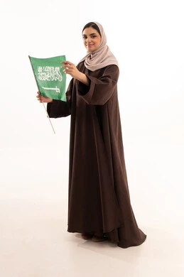 Celebrating national occasions, pride and honor in belonging to the homeland, commemorating Saudi National Day on September 23, Flag Day on March 11, raising the banner of monotheism and Islam, a portrait of a Saudi Arabian Gulf woman in a hijab holding the Saudi flag in her hand, full body length, white background.