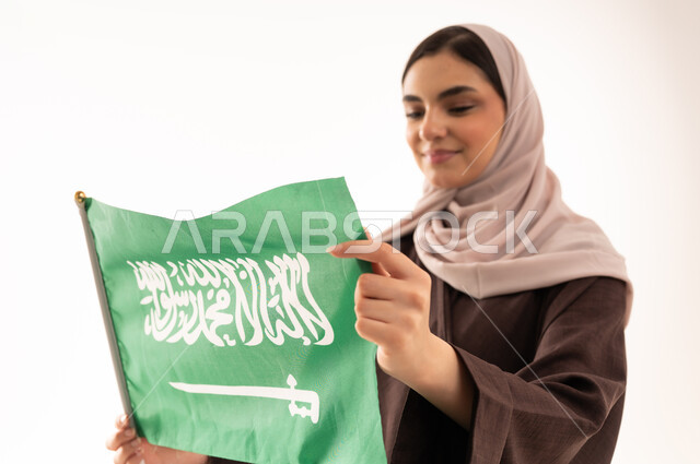 الفخر بإنجازات البلاد، رفع راية التوحيد والإسلام، الاحتفال بالمناسبات الوطنية،  الاعتزاز بالانتماء للوطن، استذكار اليوم الوطني السعودي 23 سبتمبر، يوم العلم 11 مارس، بورتريه مقرب لامرأة عربية خليجية سعودية محجبة تحمل بيدها علم السعودية ، خلفية بيضاء