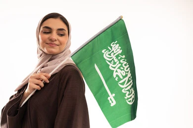 الفخر بإنجازات البلاد، رفع راية التوحيد والإسلام، الاحتفال بالمناسبات الوطنية،  الاعتزاز بالانتماء للوطن، استذكار اليوم الوطني السعودي 23 سبتمبر، يوم العلم 11 مارس، بورتريه مقرب لامرأة عربية خليجية سعودية محجبة تحمل بيدها علم السعودية ، خلفية بيضاء