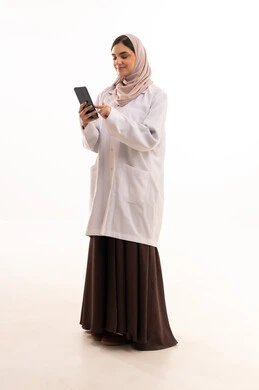 تصفح مواقع التواصل الاجتماعي عبرالهاتف المحمول ،تقديم الخدمات العلاجية في المراكز الطبية والمستشفيات،مفهوم الطب والرعاية الصحية، بورتريه لطبيبة عربية خليجية سعودية محجبة ترتدي عباءه ملونة مع المعطف الطبي تقوم بتدوين وتسجيل معلومات المرضى,  صورة الجسم بالطول الكامل، خلفيه بيضاء