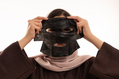 خطوات تنظيف وتقشير البشرة، مفهوم العناية والاهتمام بالمظهر الخارجي عند النساء،استخدام مكون  ومستحضر تجميلي،  بورتريه مقرب لوجه  امرأة عربية خليجية سعودية ترتدي العباءة تقوم بوضع قناع الطين على وجهها، صورة الجسم بالكامل، خلفية بيضاء