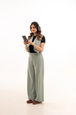 مفهوم الإصغاء والإستماع، إستخدام تقنيات الهيدفون و الهاتف المحمول ،قضاء اوقات ممتعة ومسلية،  بورتريه لشابة عربية خليجية سعودية ترتدي الزي الكاجوال  تضع سماعات الرأس الصوتية تستمع إلى الموسيقى بتعابير السعادة والسرور، صورة لكامل الجسم، خلفية بيضاء