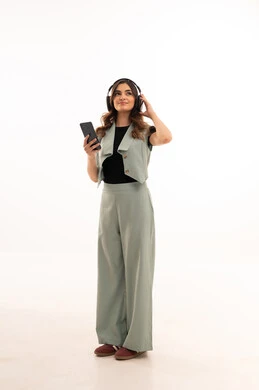 قضاء اوقات ممتعة ومسلية، مفهوم الإصغاء والإستماع، إستخدام تقنيات الهيدفون و الهاتف المحمول ، بورتريه لشابة عربية خليجية سعودية ترتدي الزي الكاجوال  تضع سماعات الرأس الصوتية تستمع إلى الموسيقى بتعابير السعادة والسرور، صورة لكامل الجسم، خلفية بيضاء