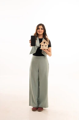 A display of a blank black screen on a mobile phone, the pursuit of owning a new house, buying and selling real estate, the pursuit of owning a new house, a portrait of a young Arab Gulf Saudi woman wearing casual attire and holding a small wooden model of a house with gestures of joy and hope, white background.