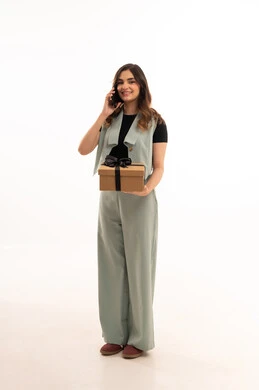 التعبير عن المحبة والتقدير,الاحتفال بأعياد الميلاد, تقديم الهدايا في المناسبات السعيدة,  بورتريه لامرأة عربية خليجية سعودية  ترتدي اللباس الكاجوال  تمسك بيديها علبة هداية و هاتف محمول لعمل مكالمة ما , شابة سعودية تنظر إلى الكاميرا بإيماءات السعادة والسرور, الجسم بالطول الكامل، خلفية بيضاء