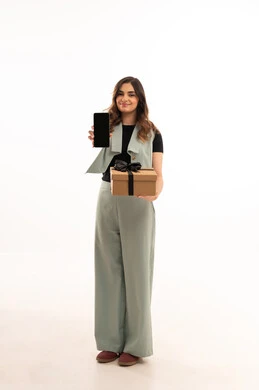 عرض شاشة سوداء  عبر الجوال ،التعبير عن المحبة والتقدير,الاحتفال بأعياد الميلاد, تقديم الهدايا في المناسبات السعيدة,  بورتريه لامرأة عربية خليجية سعودية  ترتدي اللباس الكاجوال  تمسك بيديها علبة هدية, الجسم بالطول الكامل، خلفية بيضاء