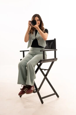 استكشاف العالم من خلال العدسة، توثيق اللحظات وتخليدها في الذاكرة، كاميرا عالية الدقة، التقاط الصور التذكارية، بورتريه لشابة عربية خليجية سعودية ترتدي الزي الكاجوال تجلس على كرسي وتقوم بالتقاط الصور الفوتوغرافية، تقنيات التصوير المبتكرة، خلفية بيضاء.