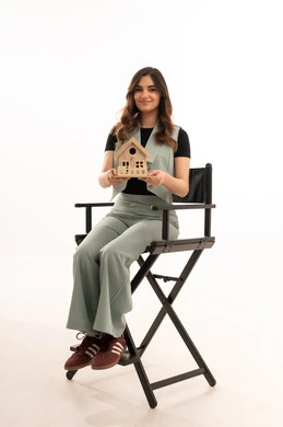 السعي لامتلاك بيت جديد، بيع وشراء العقارات، السعي لامتلاك بيت جديد، الروابط الأسرية القوية، بورتريه لشابة عربية خليجية سعودية ترتدي الزي الكاجوال تدجلس على كرسي وتحمل بيديها مجسم خشبي صغير لمنزل بإيماءات السرور والأمل, خلفية بيضاء