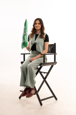 Pride in national identity, celebrating Saudi Flag Day on March 11, a portrait of a young Arab Gulf Saudi woman wearing casual attire sitting on a chair holding the Kingdom's flag with gestures of joy and happiness, commemorating the National Day for the unification of Saudi Arabia, the Saudi National Day on the 8th of Rabi' al-Awwal, white background.
