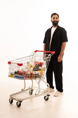 شراء المستلزمات والحاجيات الضرورية، الاستمتاع بالتسوق في المولات والمراكز التجارية، الاستفادة من الخصومات والتنزيلات على المنتجات، بورتريه لرجل عربي خليجي عماني يرتدي الزي الكاجوال يقف و عربة المشتريات أمامه ، الجسم بالطول الكامل، خلفية بيضاء