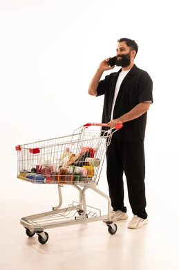 تصفح مواقع التواصل الاجتماعي ، استخدام التكنولوجيا والتقنيات الحديثة، شراء المستلزمات والحاجيات الضرورية، الاستمتاع بالتسوق في المولات والمراكز التجارية، الاستفادة من الخصومات والتنزيلات على المنتجات، بورتريه لرجل عربي خليجي عماني يرتدي الزي الكاجوال يقف و عربة المشتريات أمامه يمسك الجوال بيده ، الجسم بالطول الكامل، خلفية بيضاء