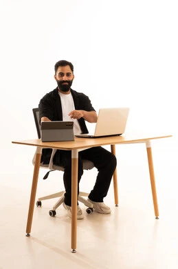 A portrait of an Arab Gulf Omani man wearing casual attire sitting on an office chair working on a laptop, conducting a live broadcast via a tablet, watching movies and series with expressions of happiness, integrating modern technologies into work, with a white background.
