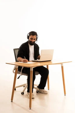 دمج التقنيات الحديثة بالمجال الإداري والمكتبي، سماع الموسيقي عبر سماعات الرأس، العمل في المكاتب والشركات العمانية، بورتريه لرجل عربي خليجي عماني يرتدي الزى الكاجوال والهيدفون يجلس على كرسي المكتب يعمل على جهاز الحاسوب المحمول يقوم بتدوين الأفكار وتسجيل المعلومات على الأوراق البيضاء، التركيز في إنجاز المهام، خلفية بيضاء