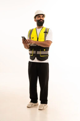 Browsing social media, using modern technologies in the engineering sector, integrating advanced technology into work, a portrait of an Arab Gulf Omani engineer wearing casual attire, a helmet, and a safety vest holding a mobile phone in his hand, full-body, white background.