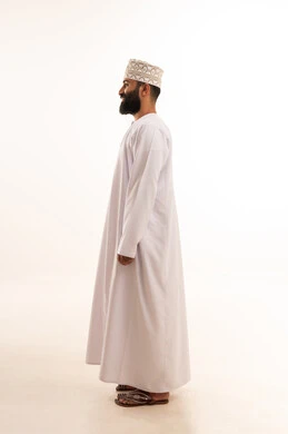 مفهوم الاناقة و الاهتمام بالمظهر الخارجي, بورتريه من الجانب لشاب عربي خليجي عماني يرتدي الدشداشة و الكمه ينظر بتعابير السعادة ، صورة الجسم بالطول الكامل, خلفية بيضاء