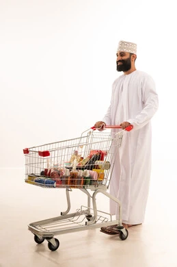 Buying essential supplies and necessities, enjoying shopping in malls and commercial centers, benefiting from discounts and sales on products, a portrait of an Arab Gulf Omani man wearing a dishdasha and kumma standing with a shopping cart in front of him, full-body, white background.