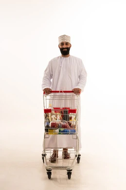 Buying essential supplies and necessities, enjoying shopping in malls and commercial centers, benefiting from discounts and sales on products, a portrait of an Arab Gulf Omani man wearing a dishdasha and kumma standing with a shopping cart in front of him, full-body, white background.