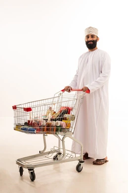 Buying essential supplies and necessities, enjoying shopping in malls and commercial centers, benefiting from discounts and sales on products, a portrait of an Arab Gulf Omani man wearing a dishdasha and kumma standing with a shopping cart in front of him, full-body, white background.