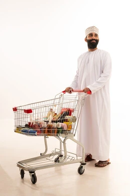 Enjoying shopping in malls and commercial centers, buying necessities and essential items, taking advantage of discounts and sales on products, a portrait of an Arab Gulf Omani man wearing a dishdasha and kumma standing with a shopping cart in front of him, full body, white background.