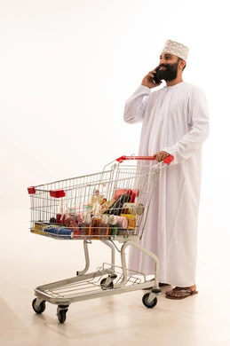 استخدام التكنولوجيا والتقنيات الحديثة، تصفح مواقع التواصل الاجتماعي ، شراء المستلزمات والحاجيات الضرورية، الاستمتاع بالتسوق في المولات والمراكز التجارية، الاستفادة من الخصومات والتنزيلات على المنتجات، بورتريه لرجل عربي خليجي عماني يرتدي الدشداشة والكمة  يقف و عربة المشتريات أمامه يمسك الجوال بيده ، الجسم بالطول الكامل، خلفية بيضاء