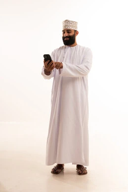 The use of modern and advanced technologies, gestures of happiness upon hearing good news, a portrait of an elderly Arab Gulf Omani man wearing a dishdasha and an Omani kumma, wearing reading glasses, holding a mobile phone in his hand and making a phone call, full-body shot, white background.